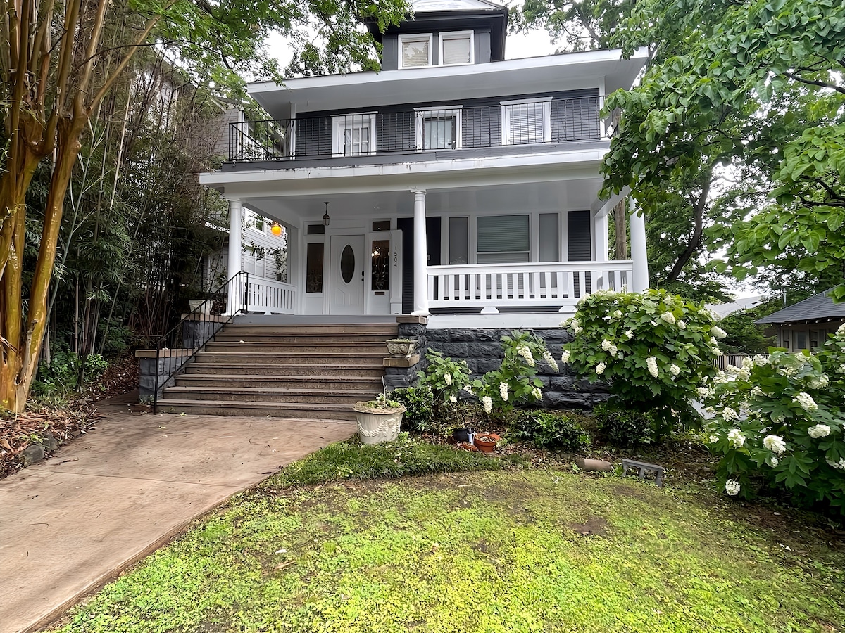 The exterior of a charming multi-story house is showcased, featuring a wide front porch with white railings and multiple large windows. Lush greenery surrounds the property, with flowering plants visible near the base of the steps leading to the entrance.