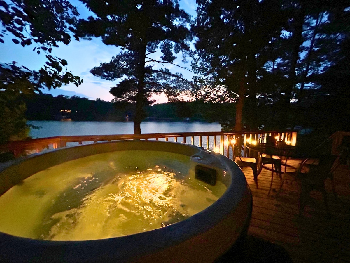 A hot tub is positioned on a wooden deck under the open sky, overlooking a tranquil lake. Surrounding trees provide privacy, while soft lights create a relaxing ambiance as the sun sets in the background.