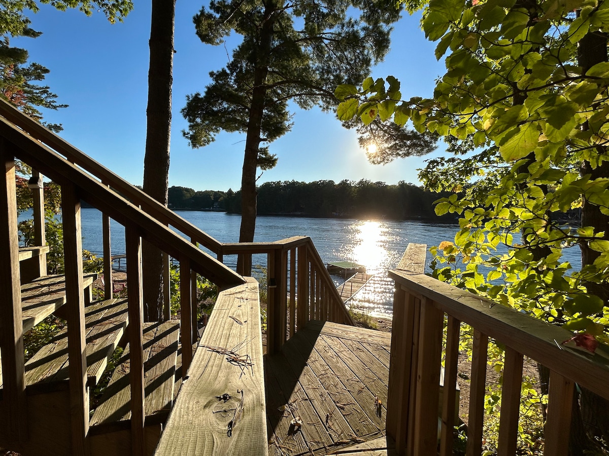 A wooden staircase leads down towards the shimmering lake, framed by lush trees and golden sunlight. Reflections on the water create a serene view, enhancing the natural surroundings and inviting relaxation by the lakeside.