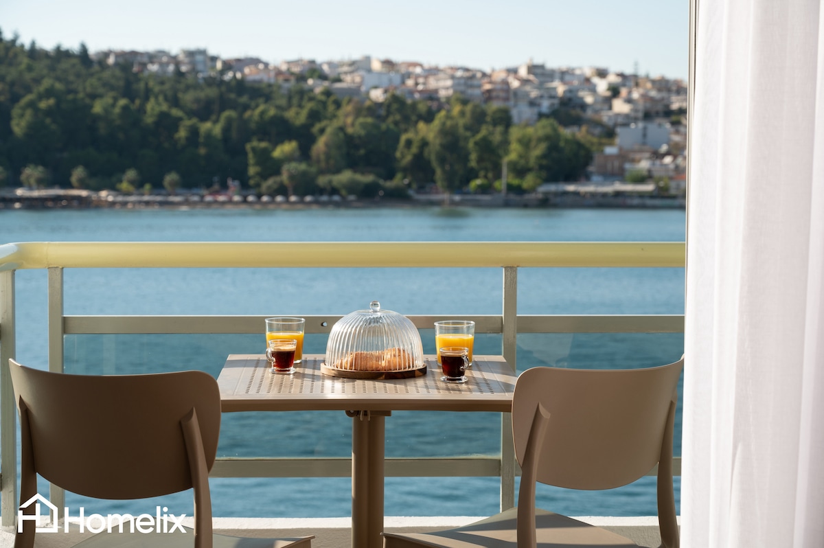 A small balcony table is set with two glasses of coffee and a covered pastry. Two chairs are positioned around the table, with a view of the serene waterfront and hills in the background. Soft natural light illuminates the scene.