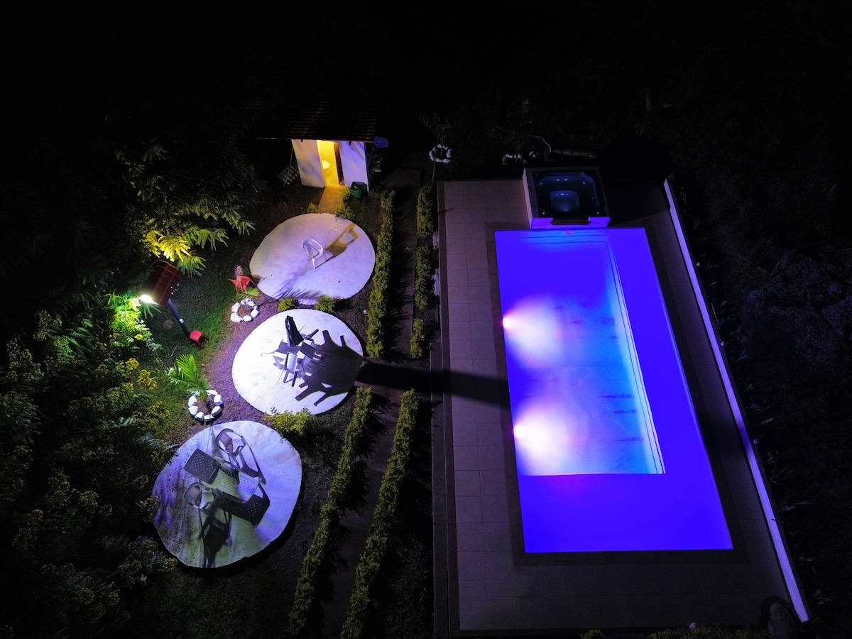 An aerial view showcases a pool illuminated in soft blue hues alongside a jacuzzi. Surrounding stone platforms with chairs feature greenery and subtle lighting, creating a serene outdoor environment ideal for relaxation.