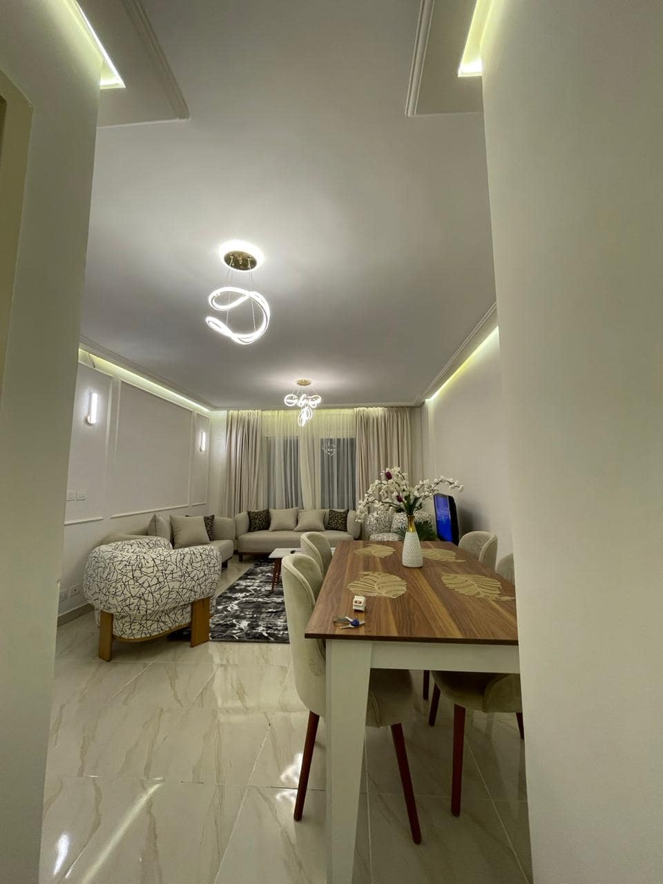 A modern living space is highlighted, featuring a sectional sofa arranged around a decorative rug. A dining table with elegant chairs sits nearby. Natural light is filtered through sheer curtains, complemented by soft overhead lighting.