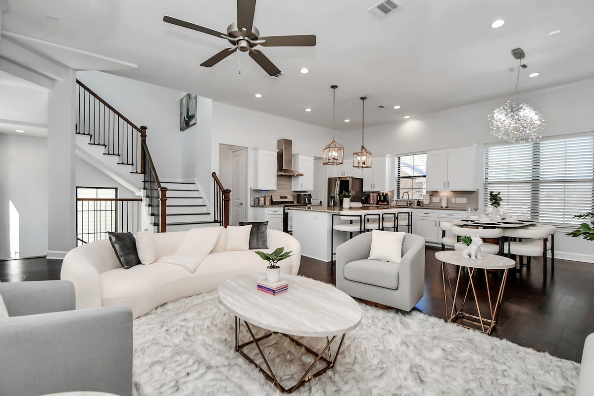 A spacious living area features a light-colored sectional sofa and two accent chairs arranged around a circular coffee table. Soft area rugs cover the dark hardwood floor. Modern lighting fixtures hang from the ceiling, and the open layout connects to a dining area and kitchen in the background.