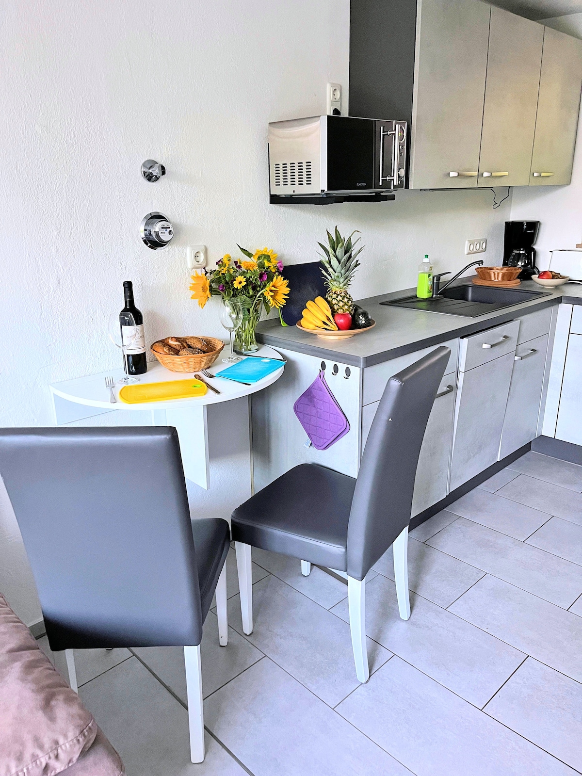 A compact kitchen area features modern cabinetry and essential appliances, including a microwave and coffee maker. A small dining table is set with two dark chairs. A bowl of fruit, a bottle of wine, and a vase with flowers add colorful accents.