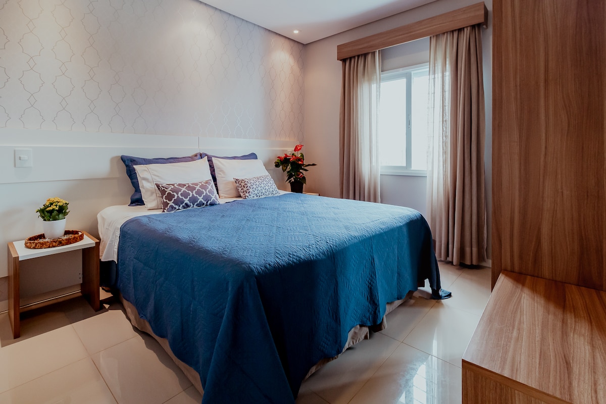 A spacious bedroom features a large bed covered with a blue quilt and several decorative pillows. Natural light enters through a window adorned with light curtains. A bedside table holds a small plant, while the wall displays an elegant pattern.