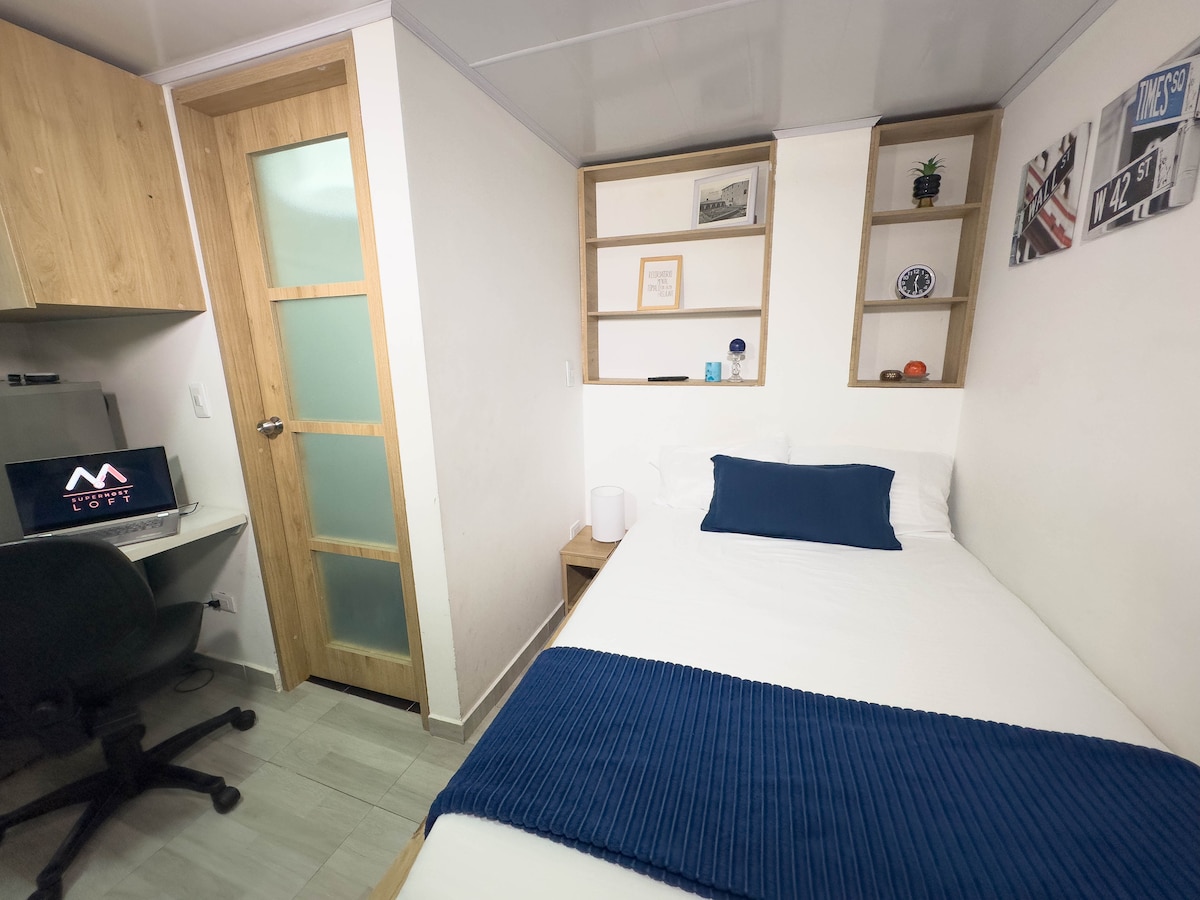 A modern studio room is captured, featuring a spacious bed with a blue throw blanket. A small desk with a computer sits in the corner, and shelves display decorative items above. A door with frosted glass leads to another area, enhancing the room's functionality.