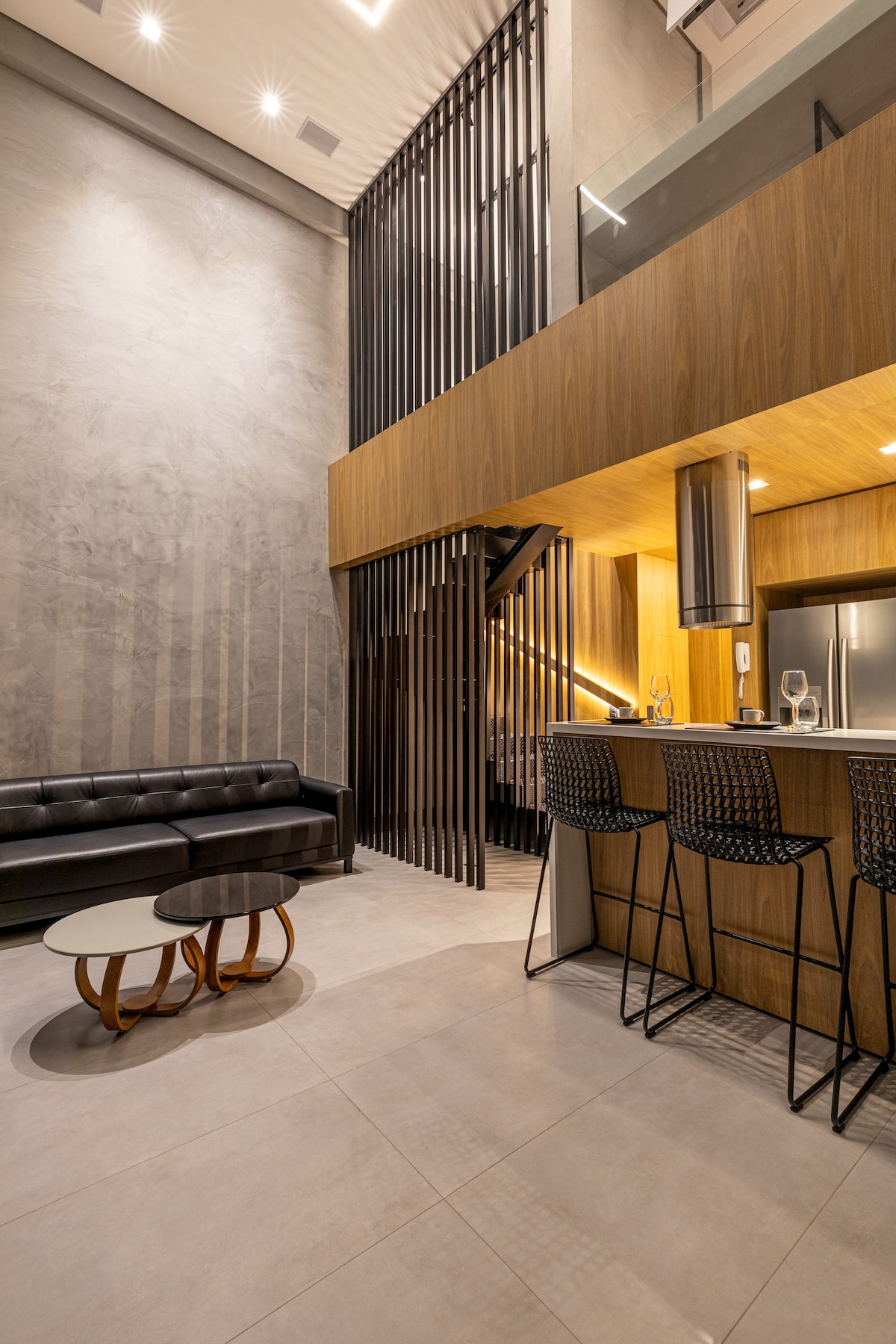 The modern loft features a double-height ceiling with a sleek wooden kitchen area. Black bar stools are positioned at the counter, complemented by a stylish black leather sofa and contemporary coffee tables. Decorative elements include textured gray walls and a striking wood and metal staircase.
