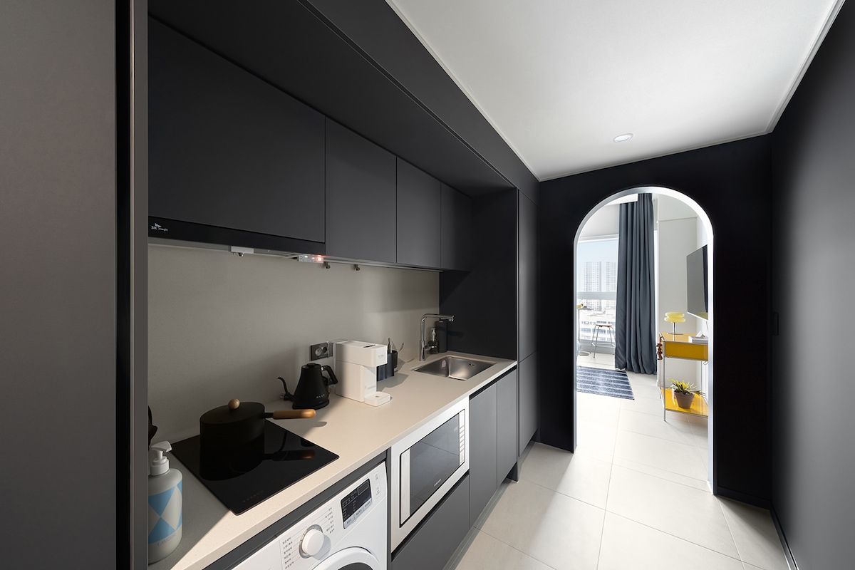 The kitchen features a modern design with dark cabinetry and a sleek countertop. A washing machine is positioned beneath a countertop, while a sink and cooking area are located alongside various appliances, including a microwave and coffee machine. An arched doorway leads to a bright living space.