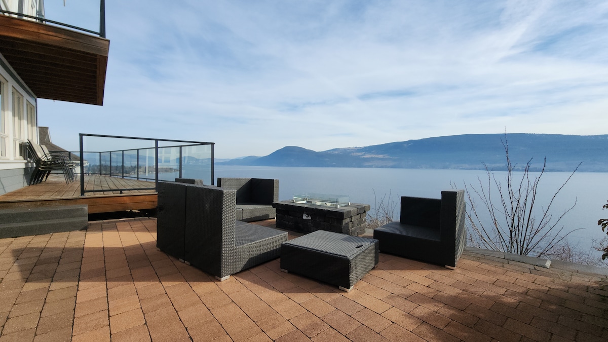 A spacious outdoor area features a paver stone patio, where modern seating arrangements surround a fire pit. Unobstructed views of the lake and rolling hills are visible in the background. The deck is accessible from the cottage, providing an inviting space for relaxation.