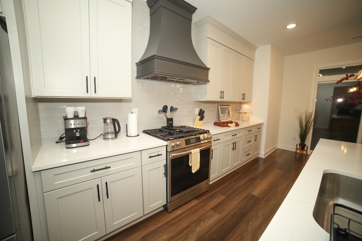 A modern kitchen showcases sleek cabinetry with a light color scheme and a large island. Stainless steel appliances, including a gas range and coffee maker, are strategically placed. A subtle backsplash complements the overall decor, while a decorative touch is provided by a framed picture and a potted plant.