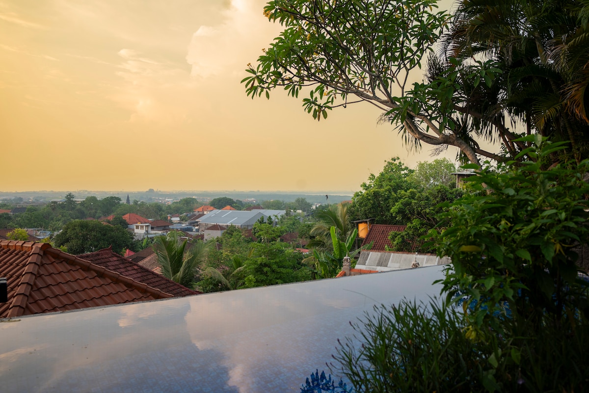 A tranquil view is presented from the edge of the infinity pool, overlooking a lush landscape with rooftops and greenery. The horizon features the soft hues of sunset blending into the sky, enhancing the serene atmosphere of the location.