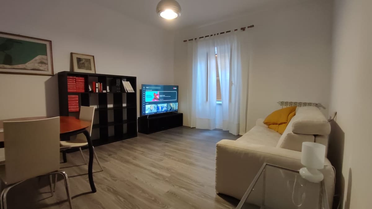 A living area is presented with light wooden flooring, featuring a comfortable sofa and a large television. A dining table with light-colored chairs is positioned nearby. Sheer white curtains frame a window, allowing natural light to illuminate the space.