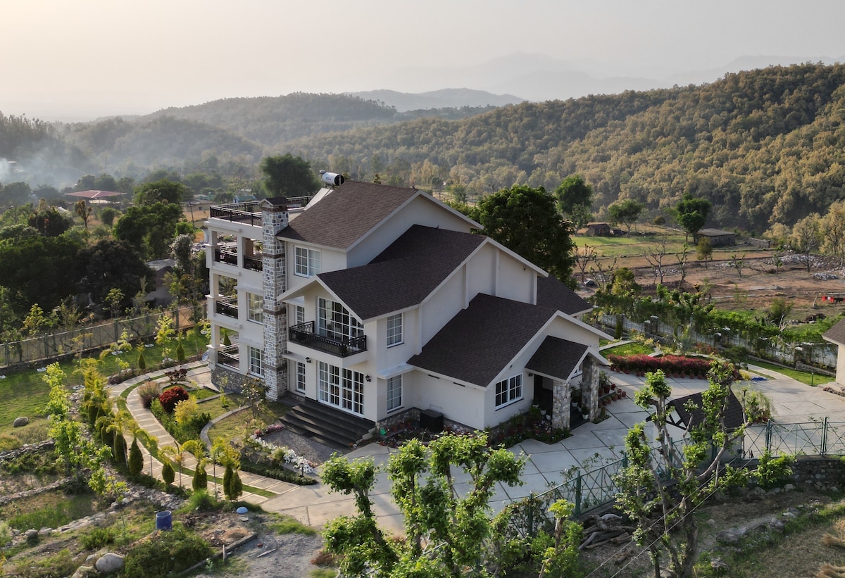 A spacious villa is set against a backdrop of rolling hills, surrounded by lush greenery. The structure showcases multiple levels, large windows, and a well-defined garden path leading to the entrance, emphasizing its inviting and tranquil setting.