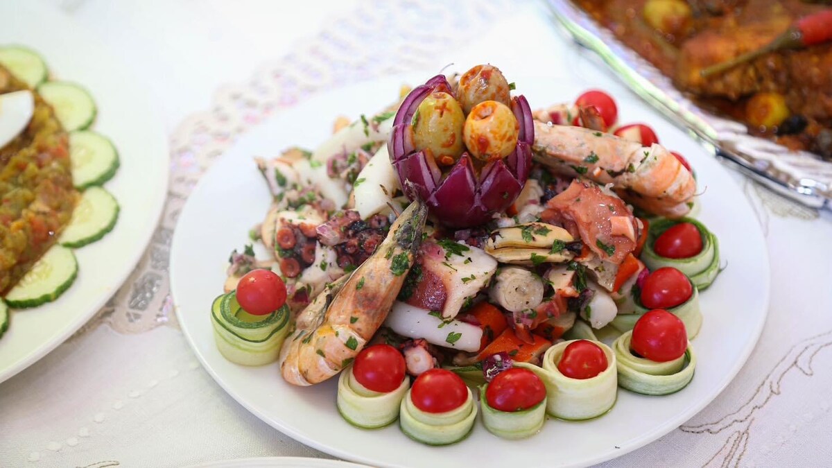 A platter of intricately arranged seafood is displayed on a white plate. The dish includes shrimp, octopus, and squid, garnished with fresh herbs. Colorful cherry tomatoes and cucumber slices accentuate the presentation, while an artfully carved vegetable adds a decorative focal point.