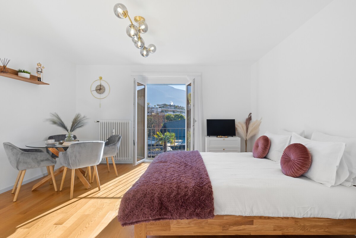 A bright and airy bedroom features a spacious bed with plush pillows and a soft blanket. A small dining area with a round table and grey chairs is visible. Large doors open to a balcony, allowing natural light to flood the space.