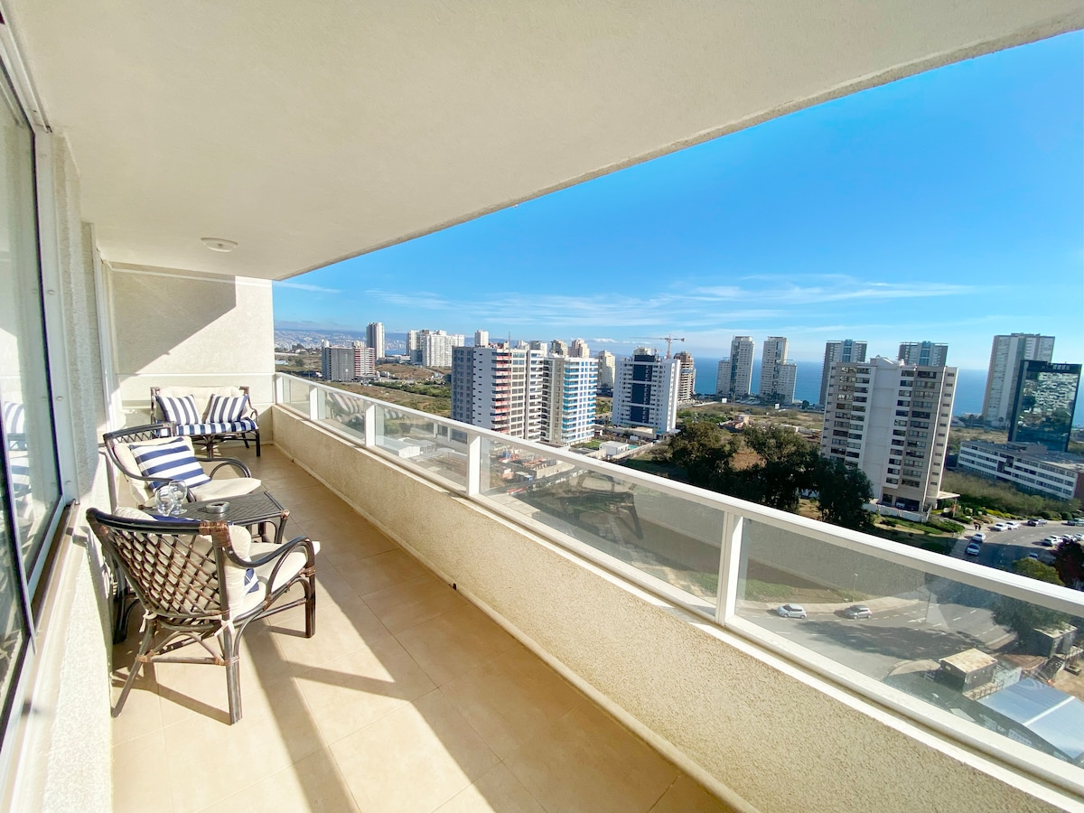 A spacious terrace extends across the unit, featuring a glass railing that offers a view of the horizon and distant buildings. Two armchairs with striped cushions are positioned next to a matching table, providing a comfortable outdoor seating area under clear blue skies.