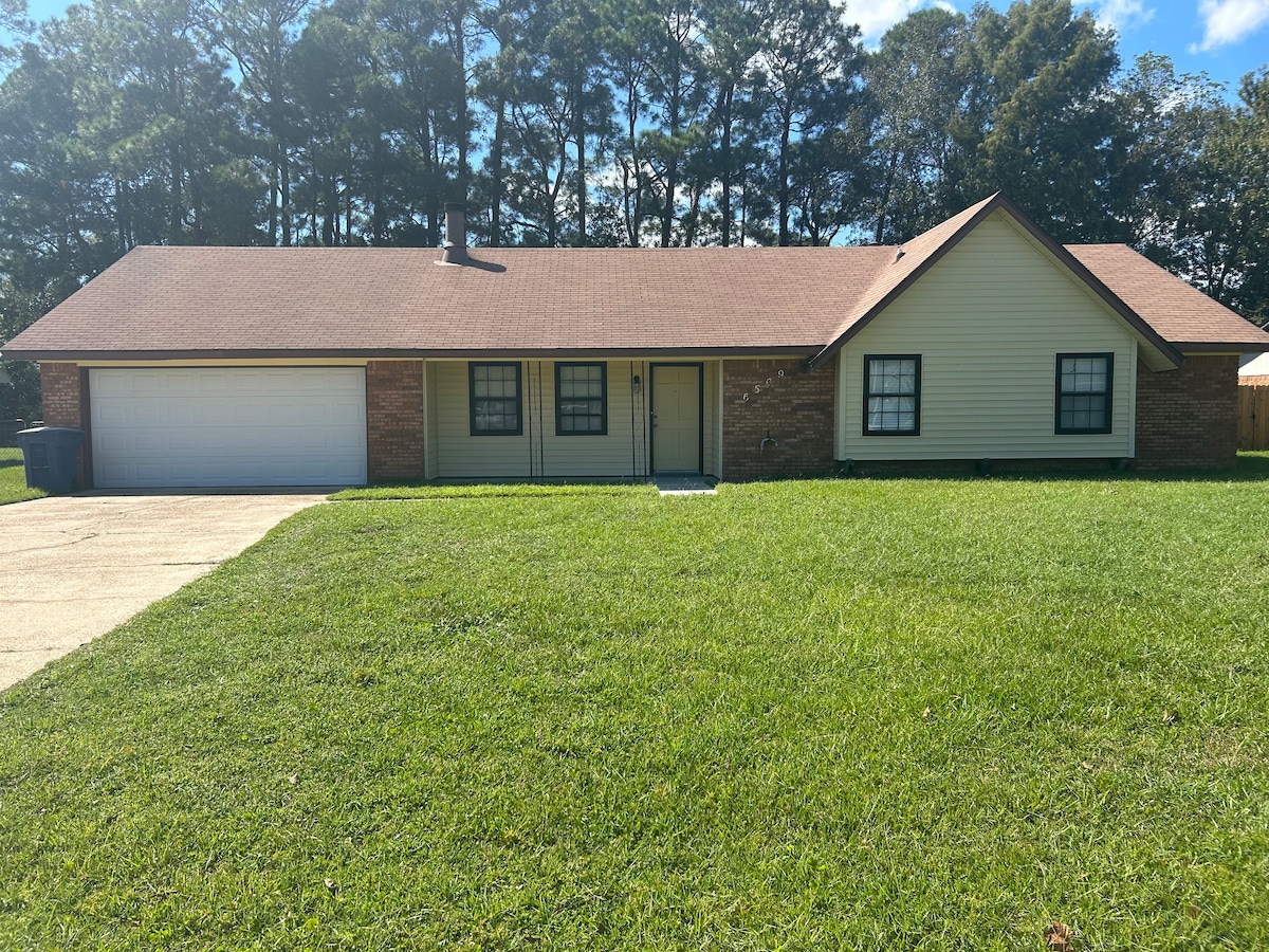 A single-story house with a brick facade and light-colored siding is surrounded by lush green grass. Large windows frame the front entrance, which is centered between the garage and the exterior. Tall trees provide natural shade in the background, enhancing the serene outdoor setting.
