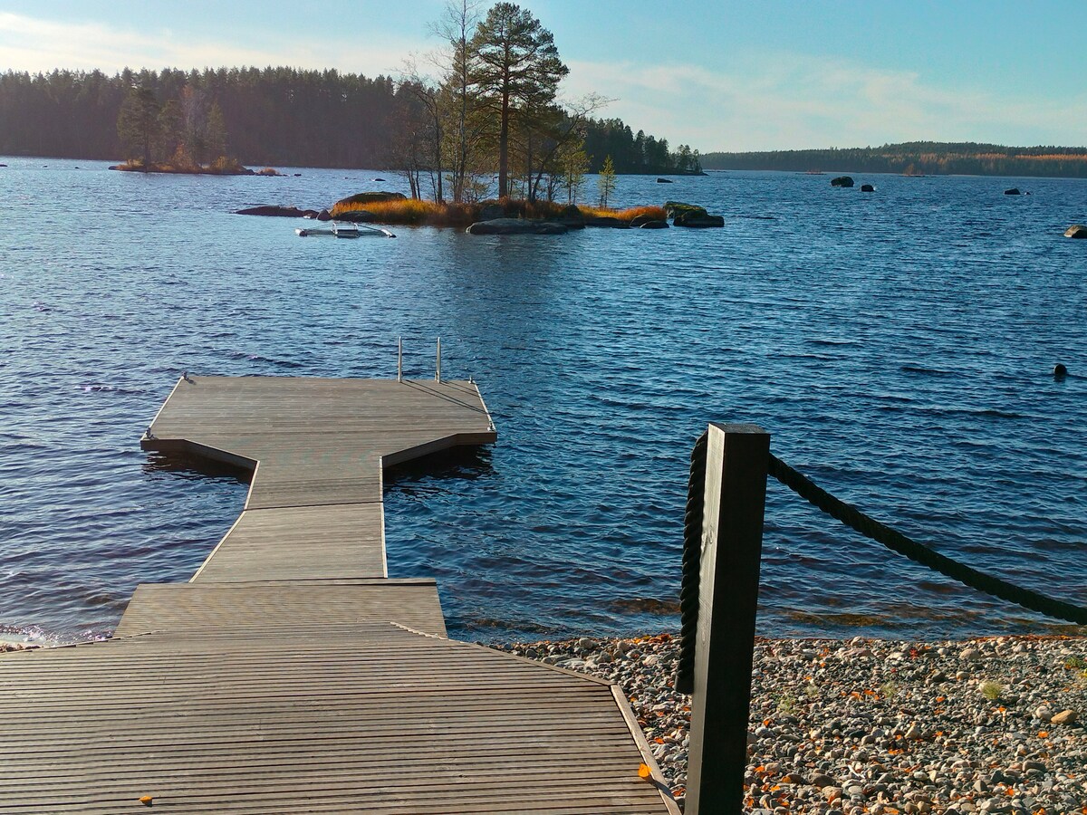 A wooden dock extends into the calm waters of Keitele Bay, leading towards small islands in the distance. Gentle waves lap against the shoreline, while trees and rocks provide a natural backdrop. The scene is illuminated by soft daylight, evoking a serene outdoor atmosphere.
