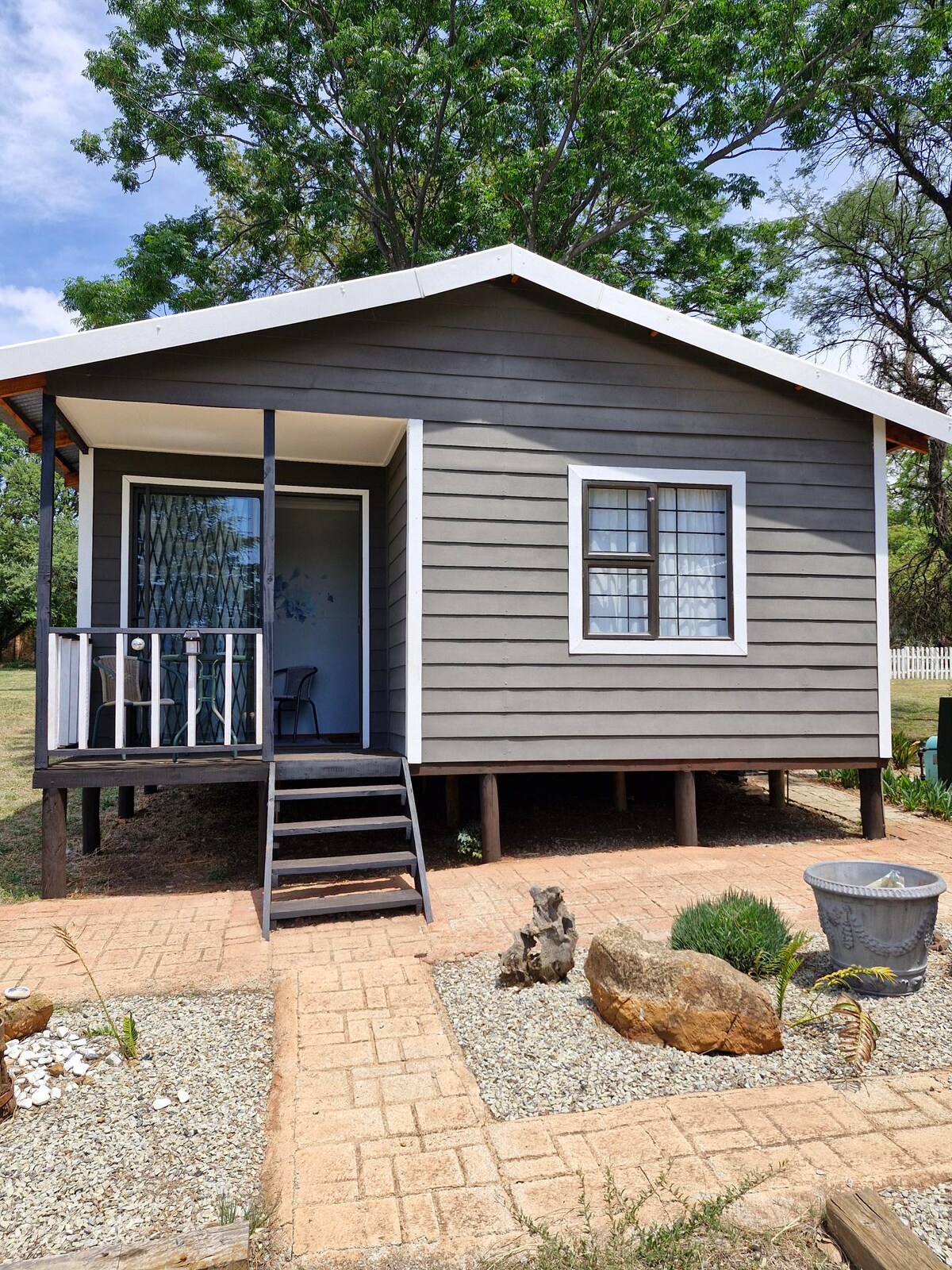 A charming cabin with a grey exterior features a small porch with steps leading up to the entrance. White window frames provide contrast against the grey walls, while a neatly arranged garden with gravel, stones, and a decorative planter enhances the outdoor space.