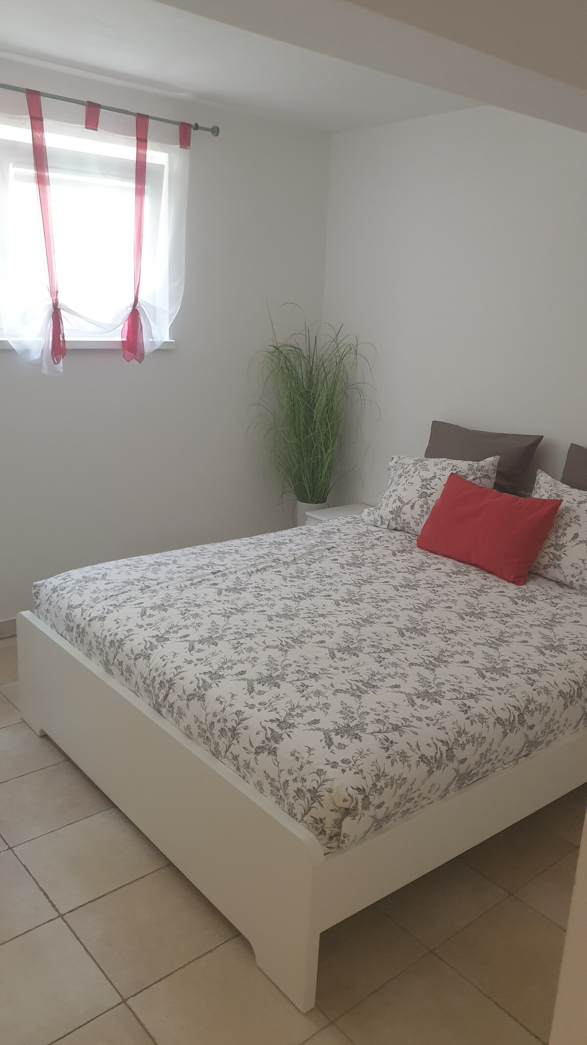 A cozy bedroom is featured, equipped with a double bed adorned with floral bedding and two decorative pillows. A small plant sits in the corner, complementing the natural light coming through the window, which is dressed with simple red and white curtains.