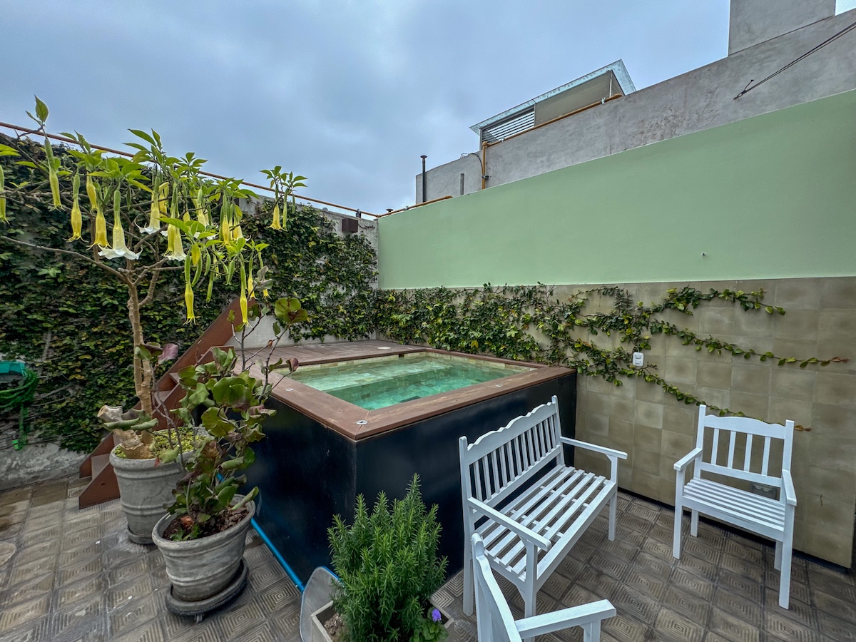 An outdoor terrace features a pool surrounded by greenery and potted plants. Two white chairs are positioned nearby, providing a space for relaxation. The area is enclosed by walls adorned with climbing plants, contributing to a serene atmosphere.
