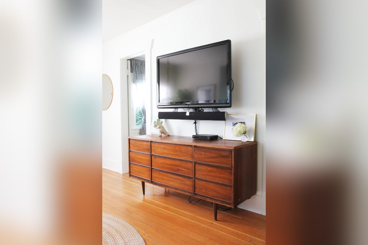 A low wooden dresser is displayed with six spacious drawers, supporting a flat-screen television. A framed photograph is positioned beside the TV, contributing to the room's functional layout. Soft natural light filters through a nearby window, enhancing the inviting atmosphere.