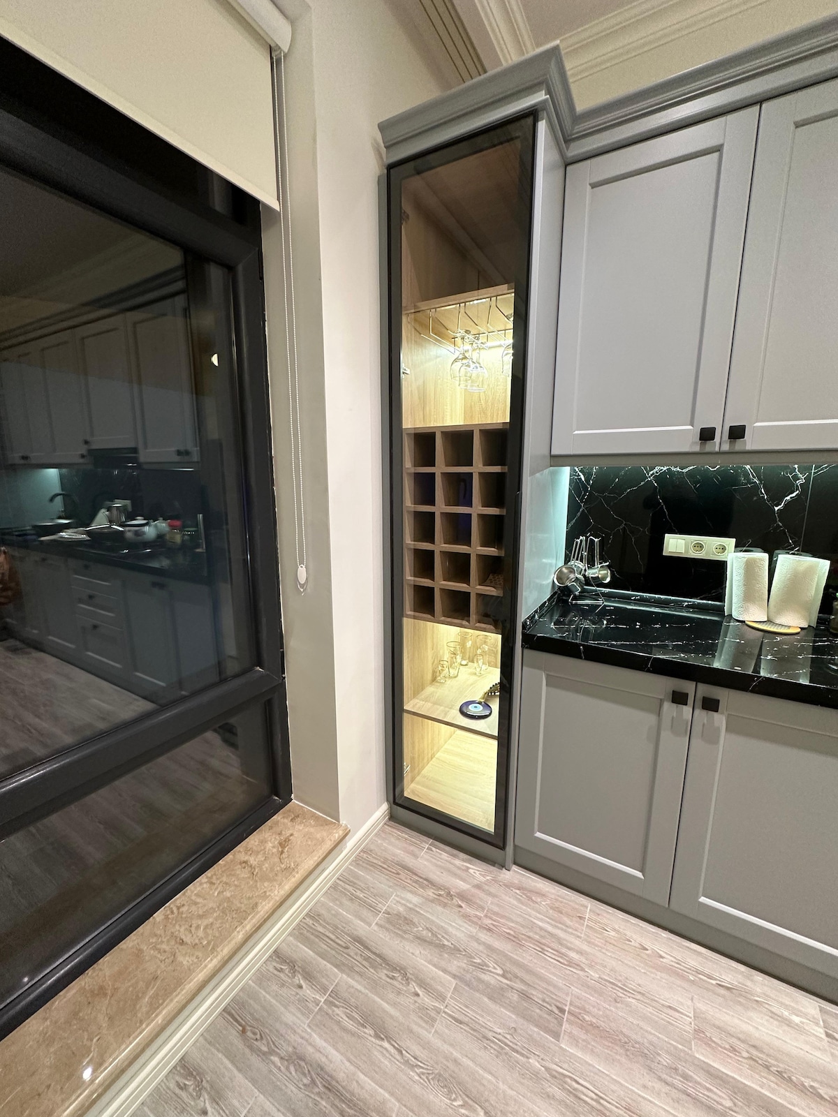 A contemporary kitchen corner features elegant cabinetry with a built-in wine rack and glass shelving, displaying various glassware. The sleek black countertop is complemented by warm lighting, creating a functional and aesthetically pleasing space. Nearby, a large window allows for natural light to enhance the ambiance.