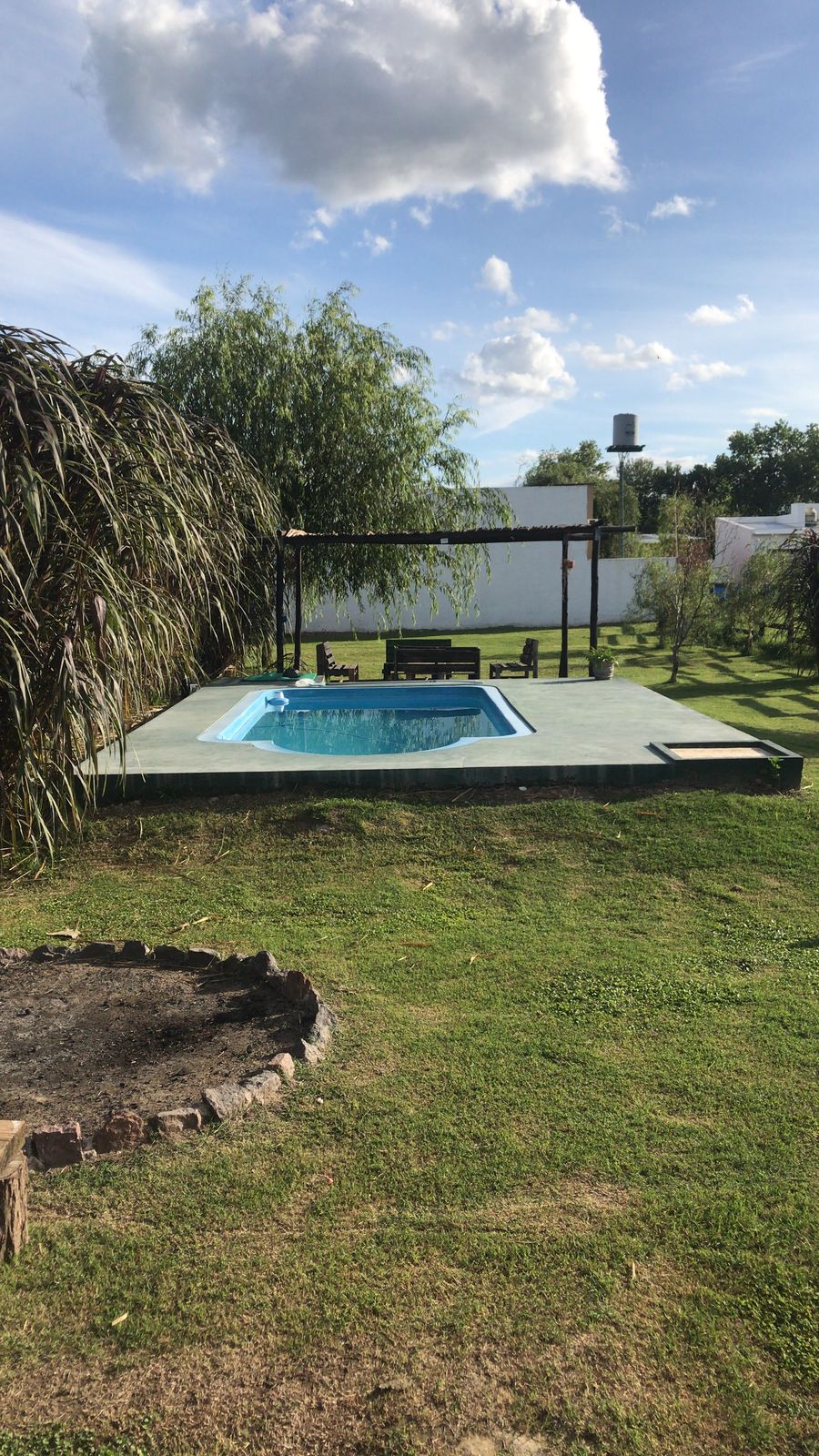 A private pool glimmers in the sunlight, set on a spacious patio surrounded by manicured grass and trees. Comfortable seating areas can be seen nearby, offering spots for relaxation. The area is enclosed by a low wall, ensuring privacy while enjoying the outdoors.