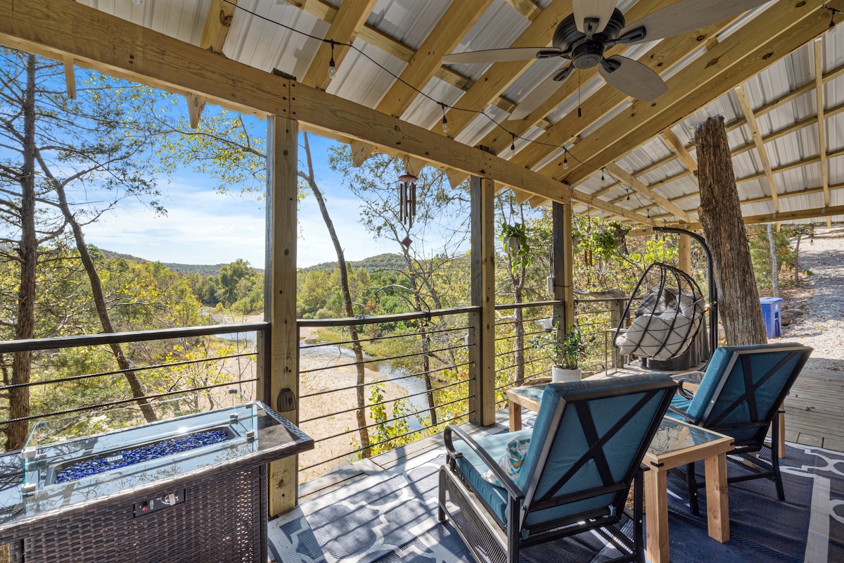 A covered outdoor deck features seating arrangements with comfortable chairs and a table. A view of the Kings River is visible beyond the railing, surrounded by lush greenery and distant hills. A ceiling fan hangs above, providing a sense of openness.