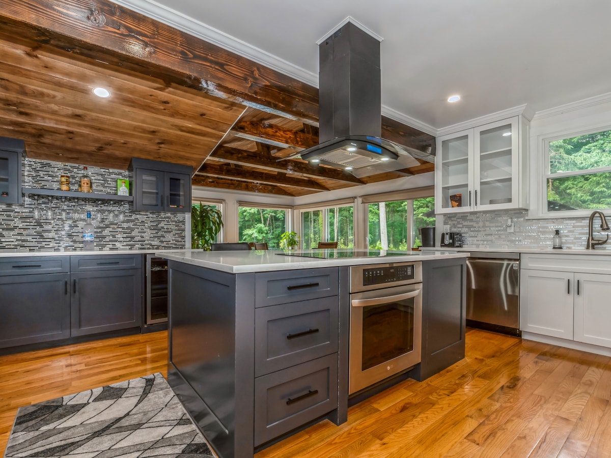 The kitchen features modern appliances, a large center island, and rich wood beams overhead. A sleek stove hood is mounted above the cooktop, with ample cabinetry for storage. Large windows provide natural light, showcasing views of the greenery outside.
