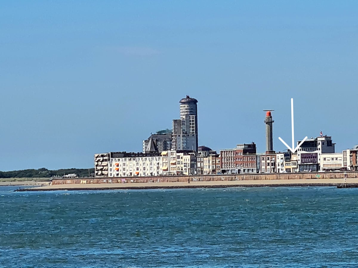 A coastal view features a series of buildings along the waterfront, showcasing a mix of contemporary and historic architecture. In the foreground, calm waters reflect sunlight, while a notable lighthouse structure stands tall among the buildings, emphasizing the seaside ambiance.