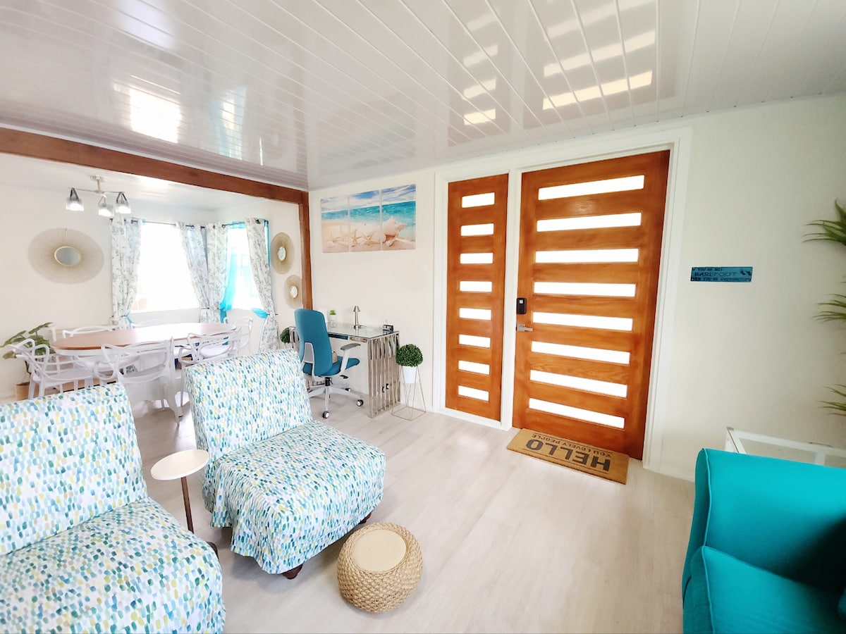 A light-filled living area features two patterned chairs positioned beside a round, woven coffee table. A welcoming front door with horizontal slats stands prominently. A dining table with several chairs is visible in the background, adorned with curtains that complement the room's coastal theme.