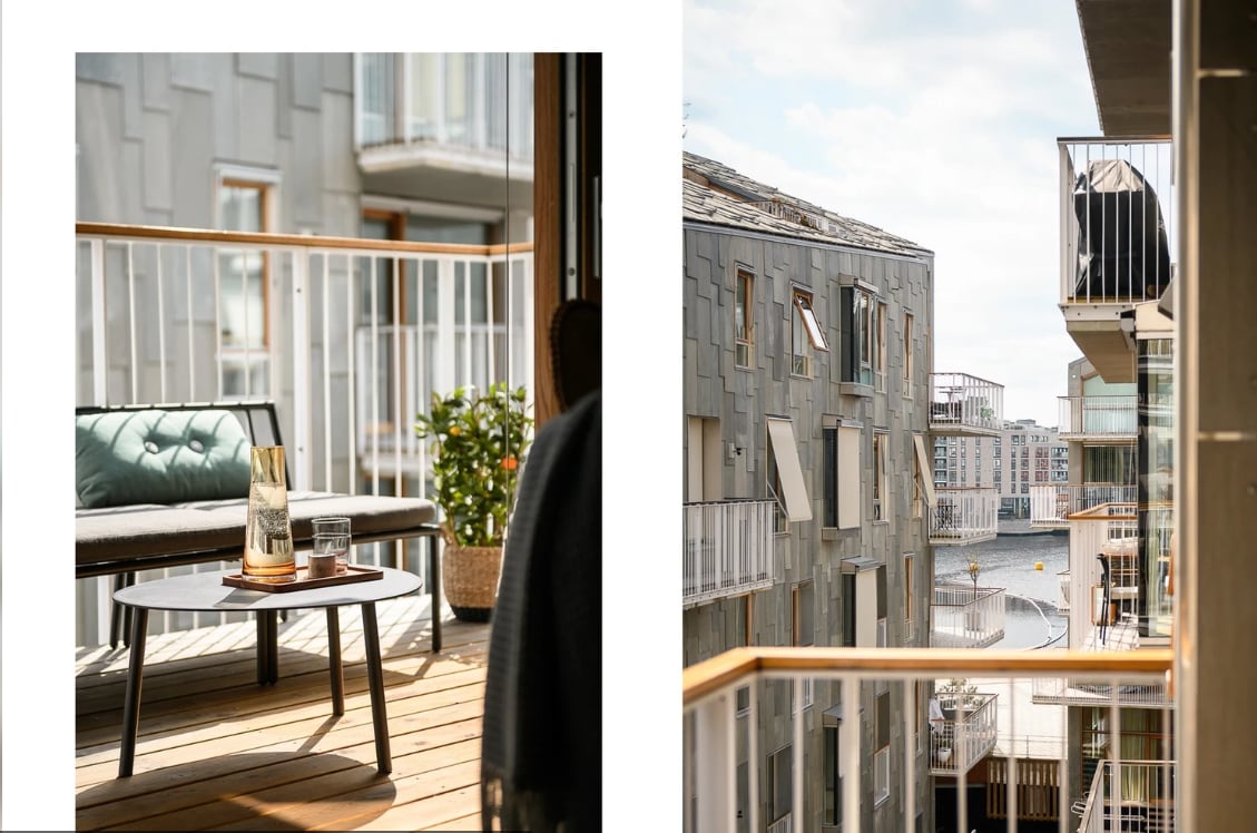 A private balcony is shown with a comfortable seating area featuring a cushioned sofa and a small round table. A decorative plant adds a touch of greenery, while the view reveals modern architecture and nearby buildings, enhancing the urban atmosphere.