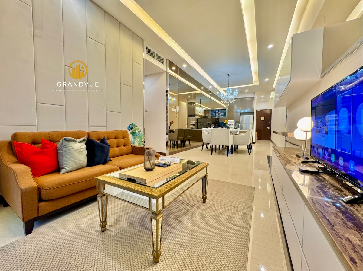 The living area features a neatly arranged brown sofa adorned with colorful cushions, positioned in front of a sleek coffee table. A modern entertainment unit displays a flat-screen TV. Dining space is visible in the background, filled with a dining table set for several guests.