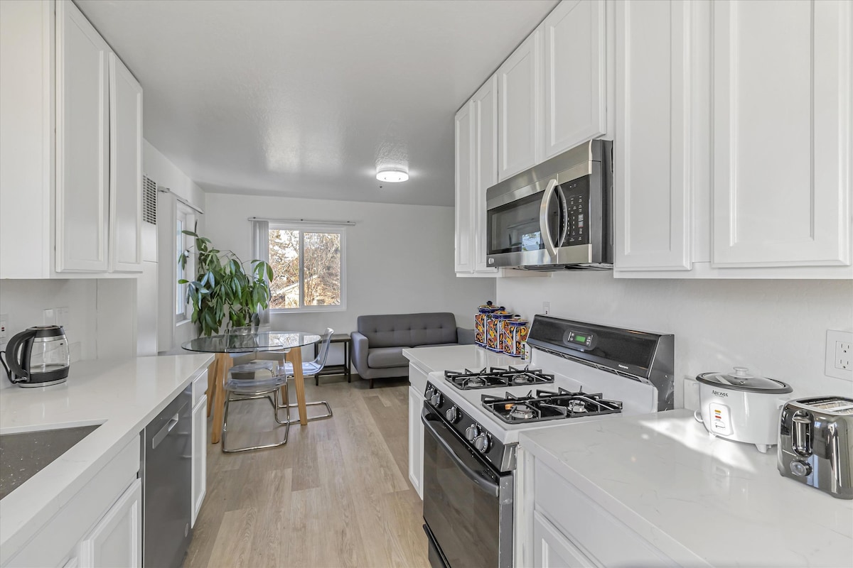 A bright kitchen area features stainless steel appliances, including a gas stove and microwave. White cabinetry is complemented by a light-colored countertop. A dining table with clear chairs is visible beside a comfortable gray sofa, and natural light flows through the window, highlighting green plants.