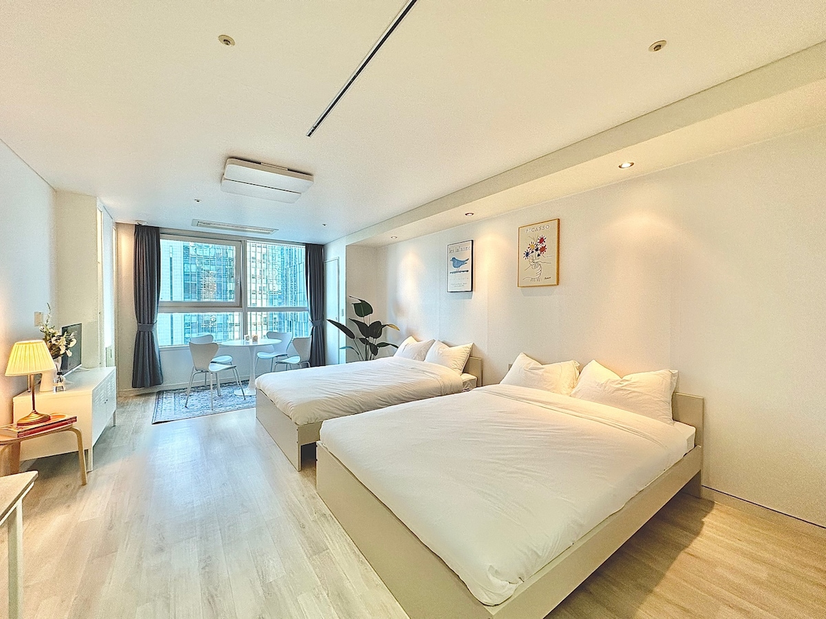 A spacious loft room features two queen beds with crisp white linens. Natural light floods the space through large windows, illuminating a simple seating area with a small table and chairs. Soft wood flooring enhances the modern decor, providing a fresh and inviting atmosphere.