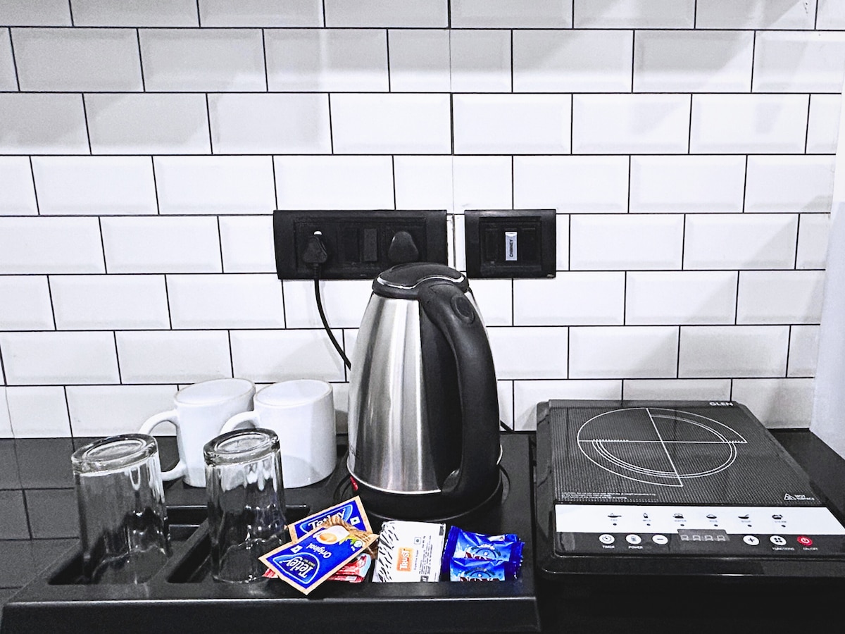 A kitchenette area features a sleek black countertop with a kettle, two clear glasses, and two white mugs. An induction cooktop is positioned beside several packets of tea, showcasing convenient amenities for light meal preparation.