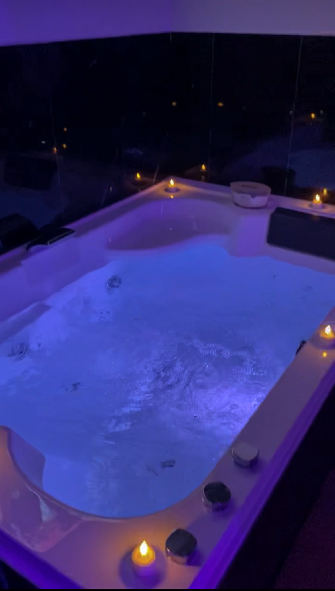 A spacious, softly lit whirlpool bath is set against dark mirrored walls. The water is illuminated by gentle purple lighting, creating a calming atmosphere. Candles are placed around the edge, enhancing the serene setting for relaxation and intimacy.