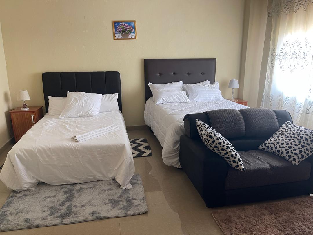 The room features two queen-sized beds dressed in white linens, complemented by bedside tables with lamps. A cozy couch with patterned cushions sits nearby on a textured rug, while pale curtains drape over a window, allowing natural light to fill the space.