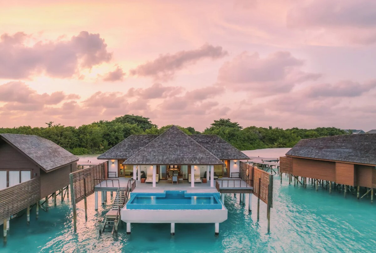 A stunning aerial view of the villa is presented, highlighting the private pool at the center. The resort is surrounded by calm waters and lush greenery, with several villas extending from the main structure. A soft sunset glow illuminates the scene, creating a serene ambiance.