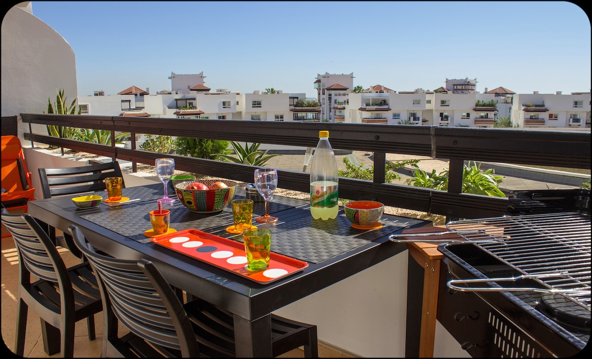 A dining table set for outdoor meals is arranged with colorful dishes and glasses, alongside a grill. Lush greenery and distant buildings are visible, framed by a wooden railing. Clear blue skies contribute to a relaxing atmosphere on the spacious terrace.