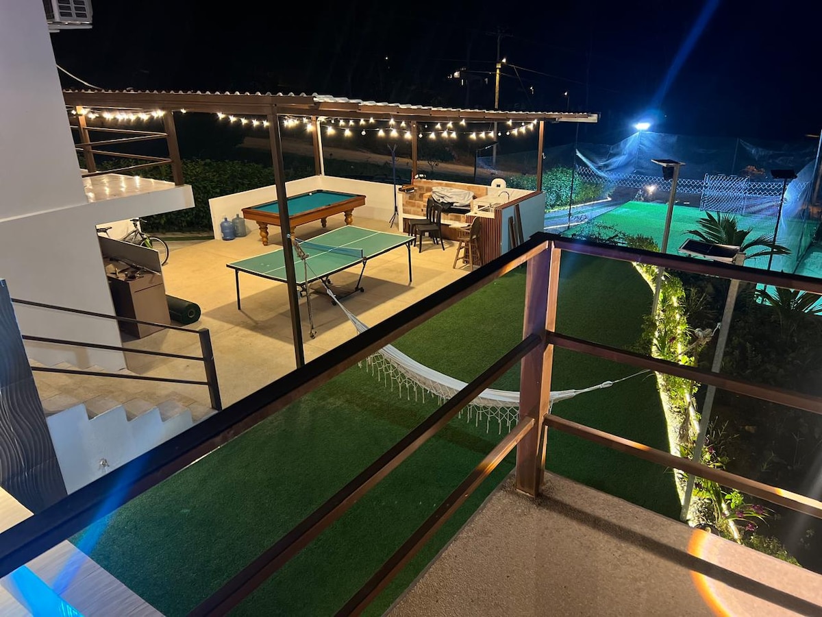 An outdoor entertainment area is visible, featuring a ping pong table and a billiards table under soft lighting. A well-maintained lawn stretches across the space, alongside a hammock. The scene is enclosed by greenery, enhancing the inviting atmosphere of the property.