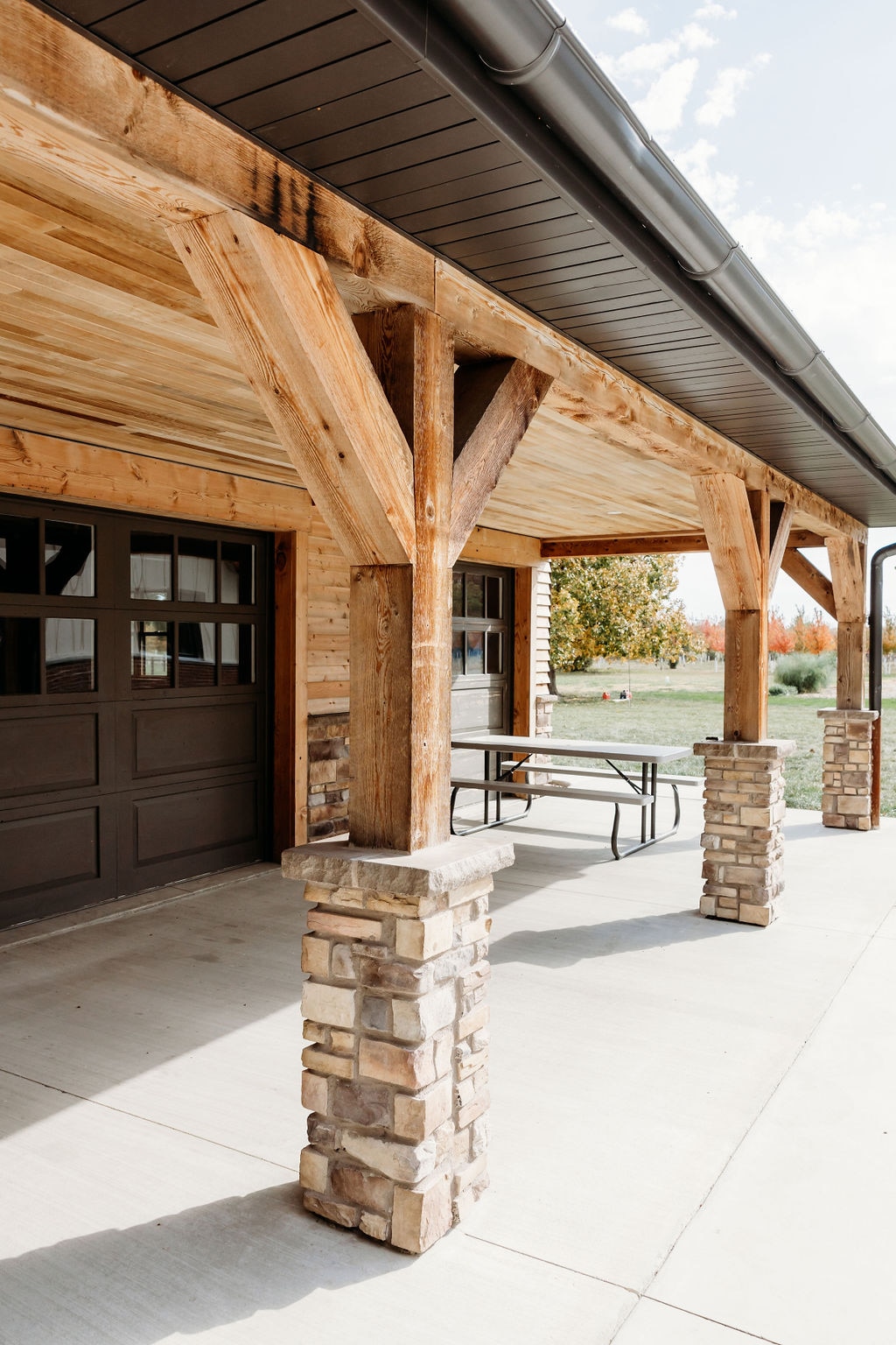 The entrance of the loft is framed by sturdy wooden beams, supported by rustic stone columns. A concrete patio leads to the entrance, creating a seamless connection to the outdoors. The surrounding landscape includes grassy areas and trees, contributing to a natural setting.