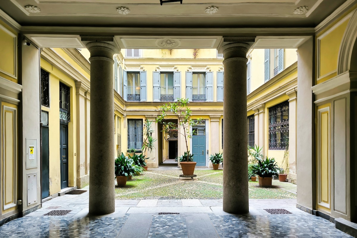 The entrance to a courtyard is framed by two columns. A central tree is surrounded by potted plants, with buildings displaying blue shutters in the background. The flooring is a mix of stone tiles and cobblestones, creating a charming atmosphere.