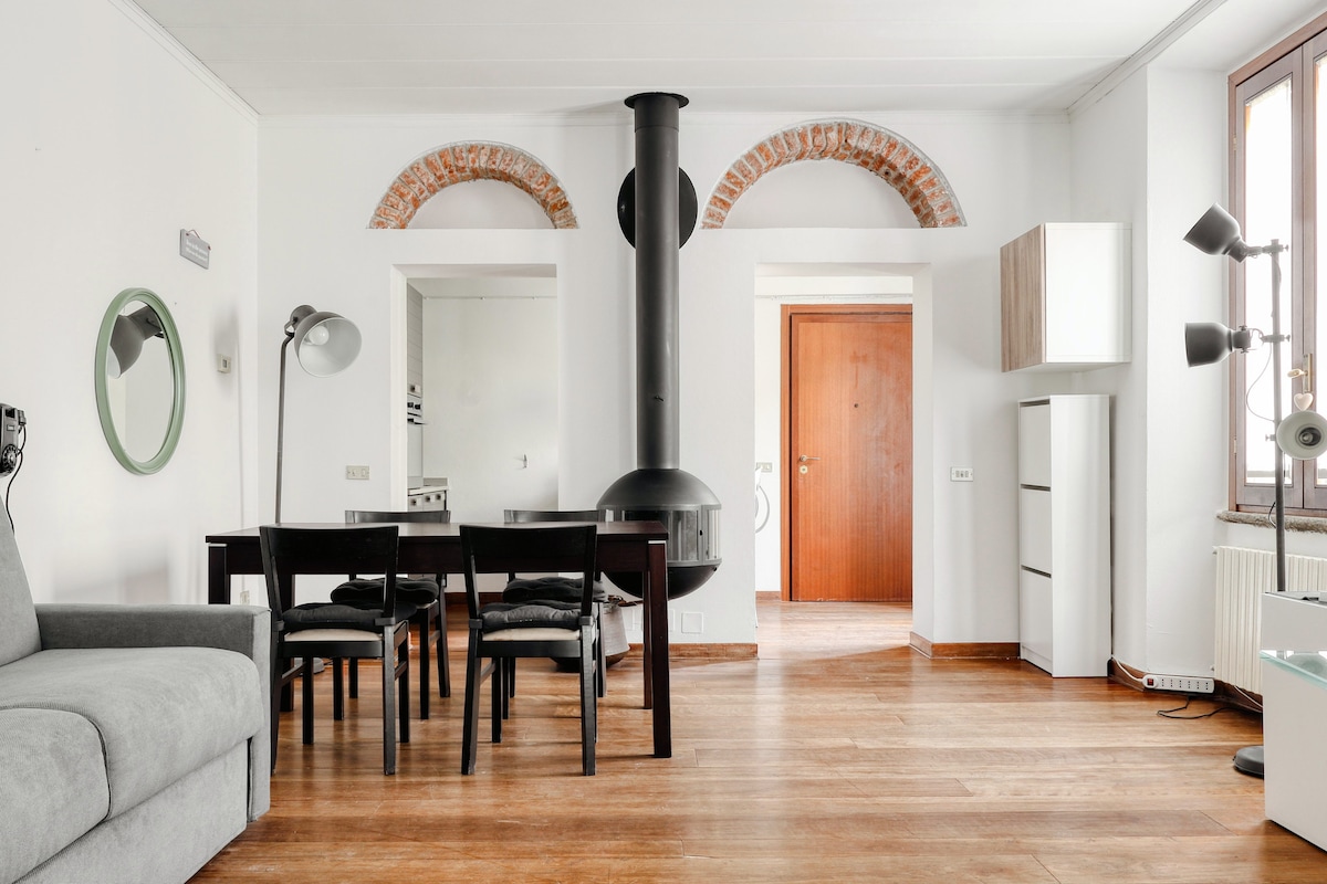 An open living area features a dining table with four black chairs, adjacent to a gray sofa. A black chimney stands prominently in the center. Natural light enters through large windows, highlighting wooden flooring and brick archways above. A door leads to a separate room.