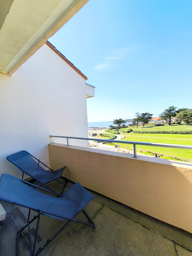 A small balcony is visible, featuring two lightweight chairs positioned for comfort. Beyond the balcony, a scenic view of a green pathway and the coastline can be seen, accentuated by a clear blue sky. The surroundings suggest a peaceful outdoor space perfect for relaxation.