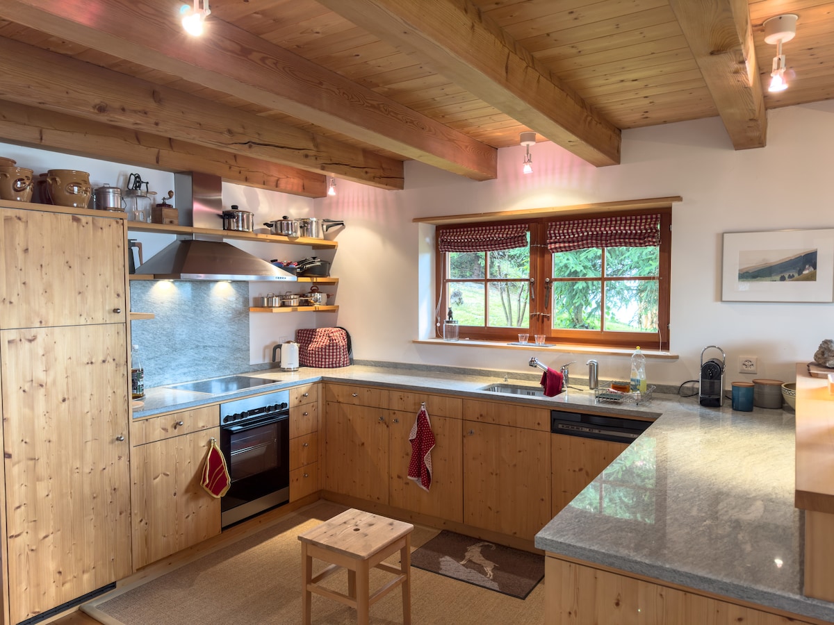 A warm kitchen space features wooden cabinetry and granite countertops. Shelves hold kitchenware, while a stove and dishwasher are placed for convenience. Large windows allow natural light to fill the area, accented by wooden beams on the ceiling.