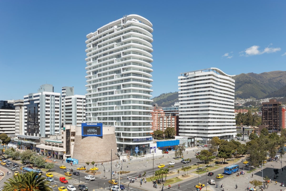 A modern high-rise building is pictured, featuring a unique curved design. The surrounding area showcases urban infrastructure with multiple vehicles and pedestrians. Nearby buildings exhibit a variety of architectural styles, while vibrant green trees add a touch of nature to the cityscape.