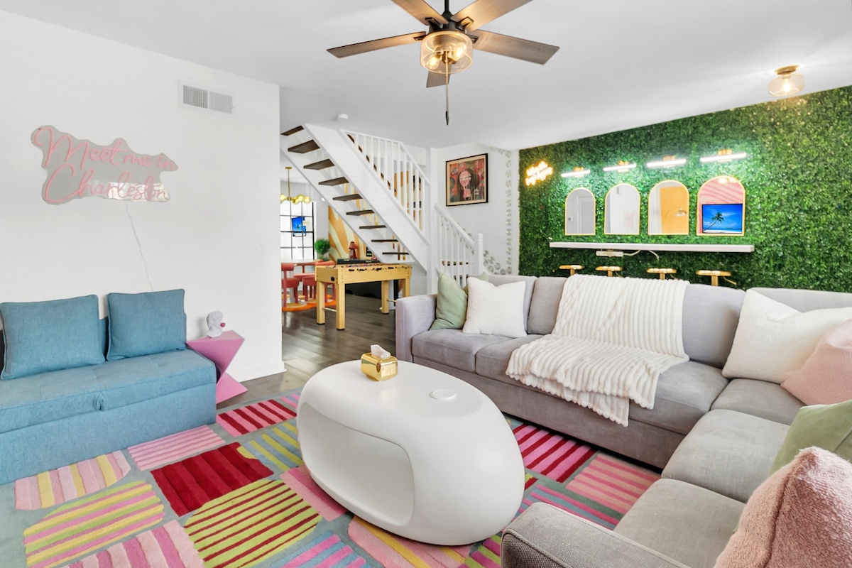 A spacious living area is highlighted by a large sectional sofa with colorful throw pillows and a vibrant, patterned rug. A whimsical wall with greenery features mounted decor and screens. A ceiling fan adds a cool element, while a staircase leads to the upper level.