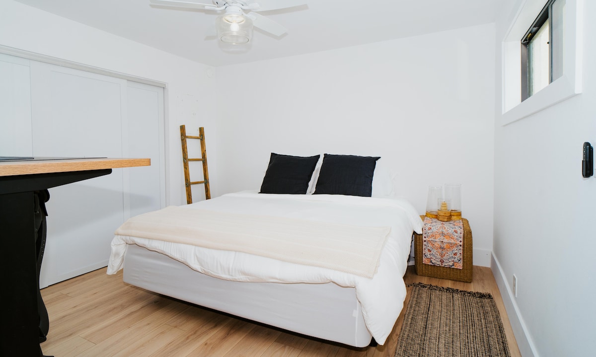 Minimalist bedroom with white linens, black pillows & woven rug — features ladder rack, patterned cloth nightstand & ceiling fan for clean, tropical comfort